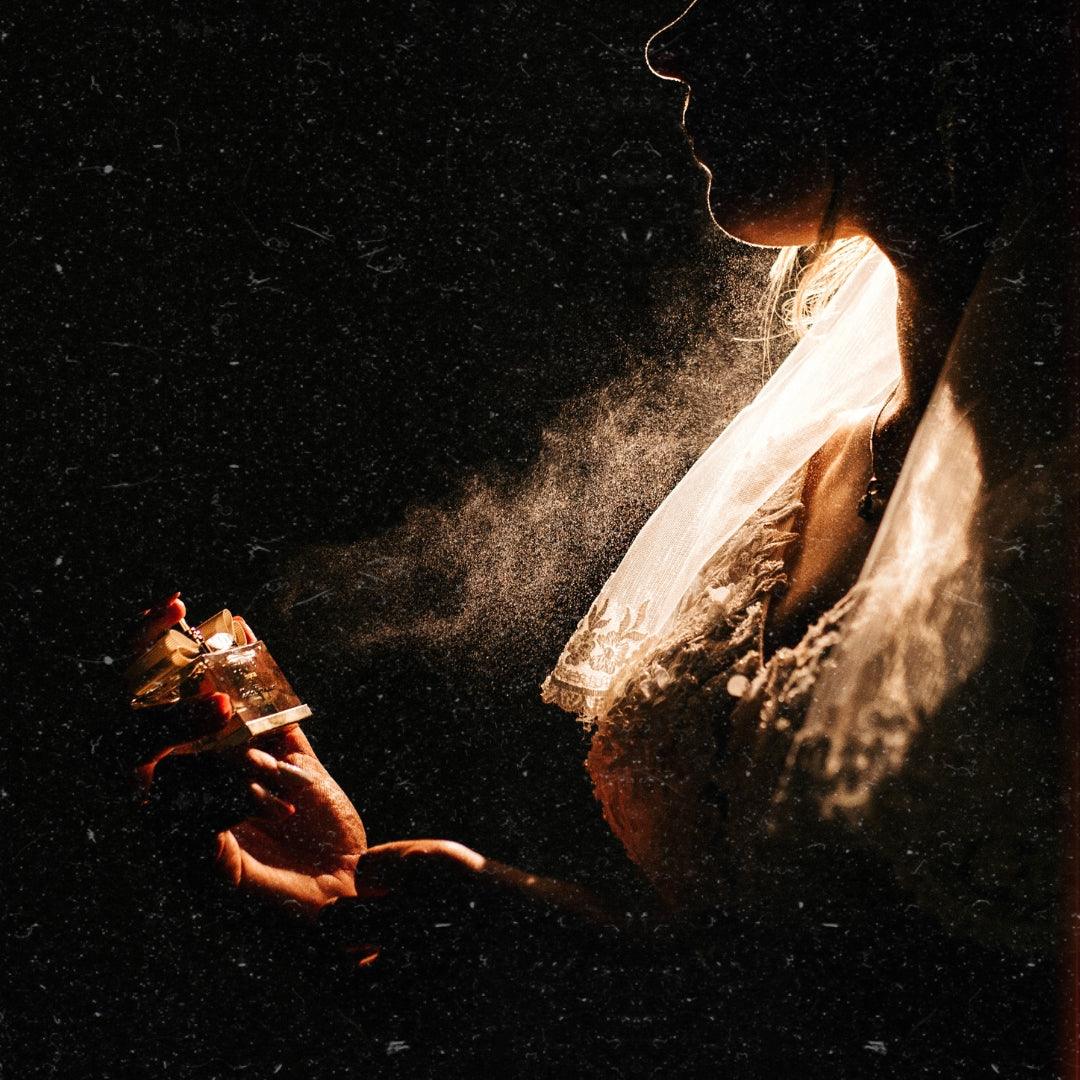 A woman elegantly sprays perfume on her neck in a dimly lit room. Soft light highlights the mist as it casts delicate shadows, creating a mysterious and intimate atmosphere