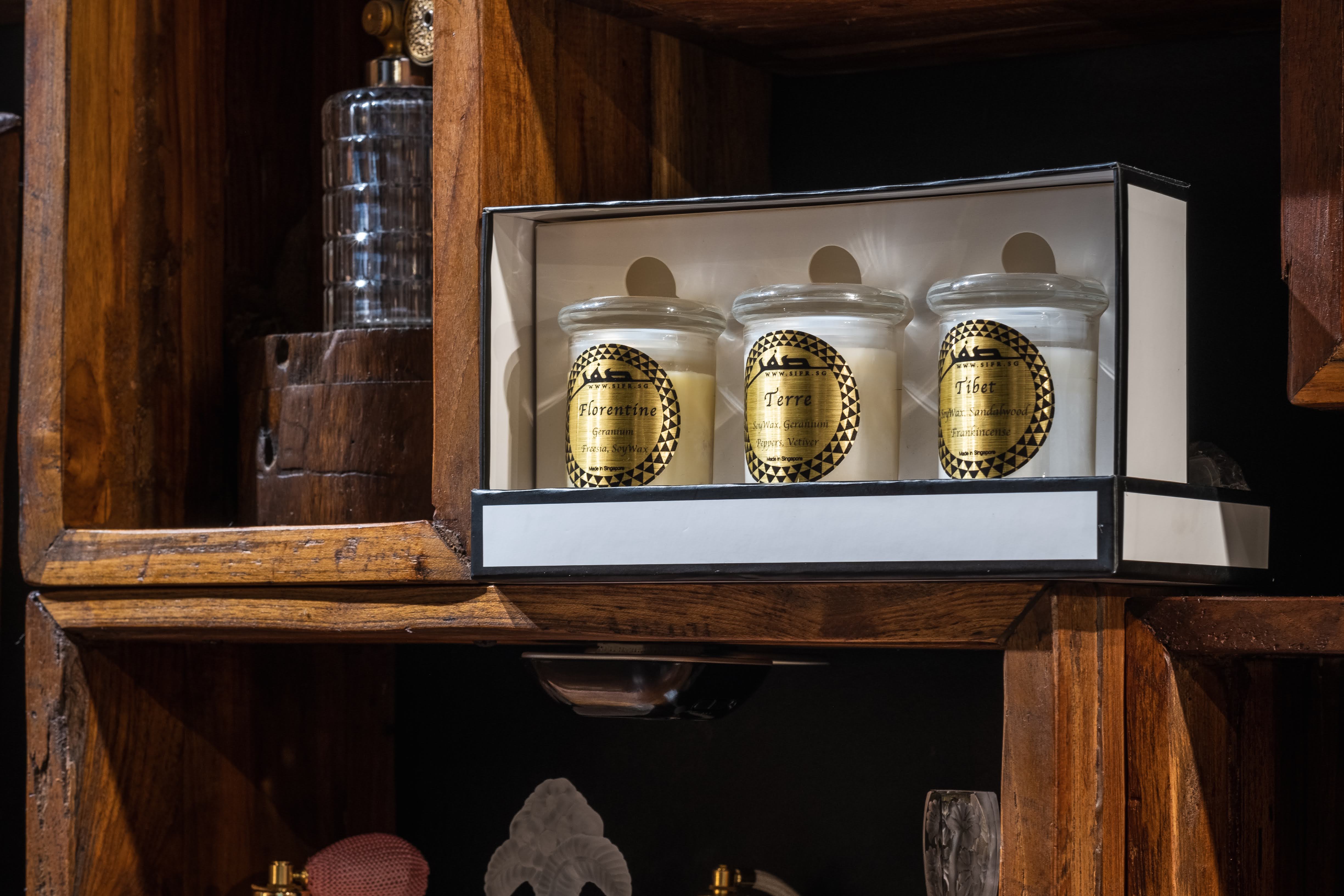 Three Sifr Aromatics soy-wax candles labeled Geranium, Peppers and Vetiver, and Frankincense, displayed in a wooden cabinet