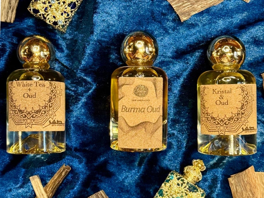 Three gold bottles labeled 'White Tea Oud', 'Burma Oud', and 'Kristol Oud' on a blue textured background.