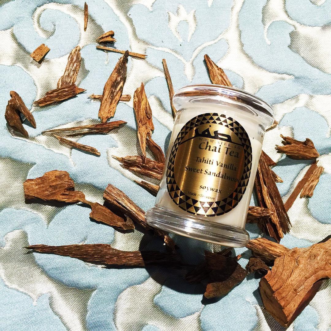 Small 'Chai Tea' candle in a white container with a glass lid, lying on a blue and white satin cloth with intricate shapes. The gold label with black text reads 'Chai Tea, Soy Wax, Black Tea, Cardamom, Oud,' and features the Sifr Aromatics logo in black. Argawood chips are placed alongside the candle