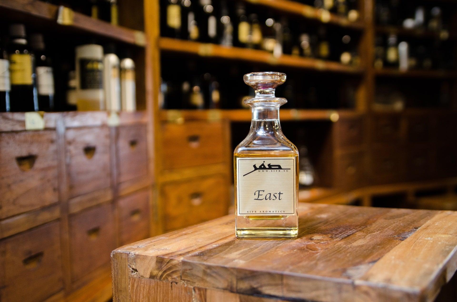 Ornate glass bottle for perfume oil with a gold label displaying the Sifr Aromatics logo and the name 'East.' The background features an out-of-focus wooden perfume organ with various ingredients, creating an aesthetically pleasing scene