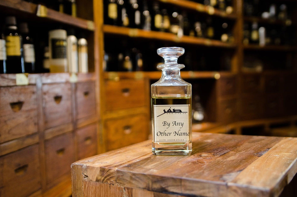 Ornate glass bottle for perfume oil with a gold label displaying the Sifr Aromatics logo and the name 'By Any Other Name.' The background features an out-of-focus wooden perfume organ with various ingredients, creating an aesthetically pleasing scene