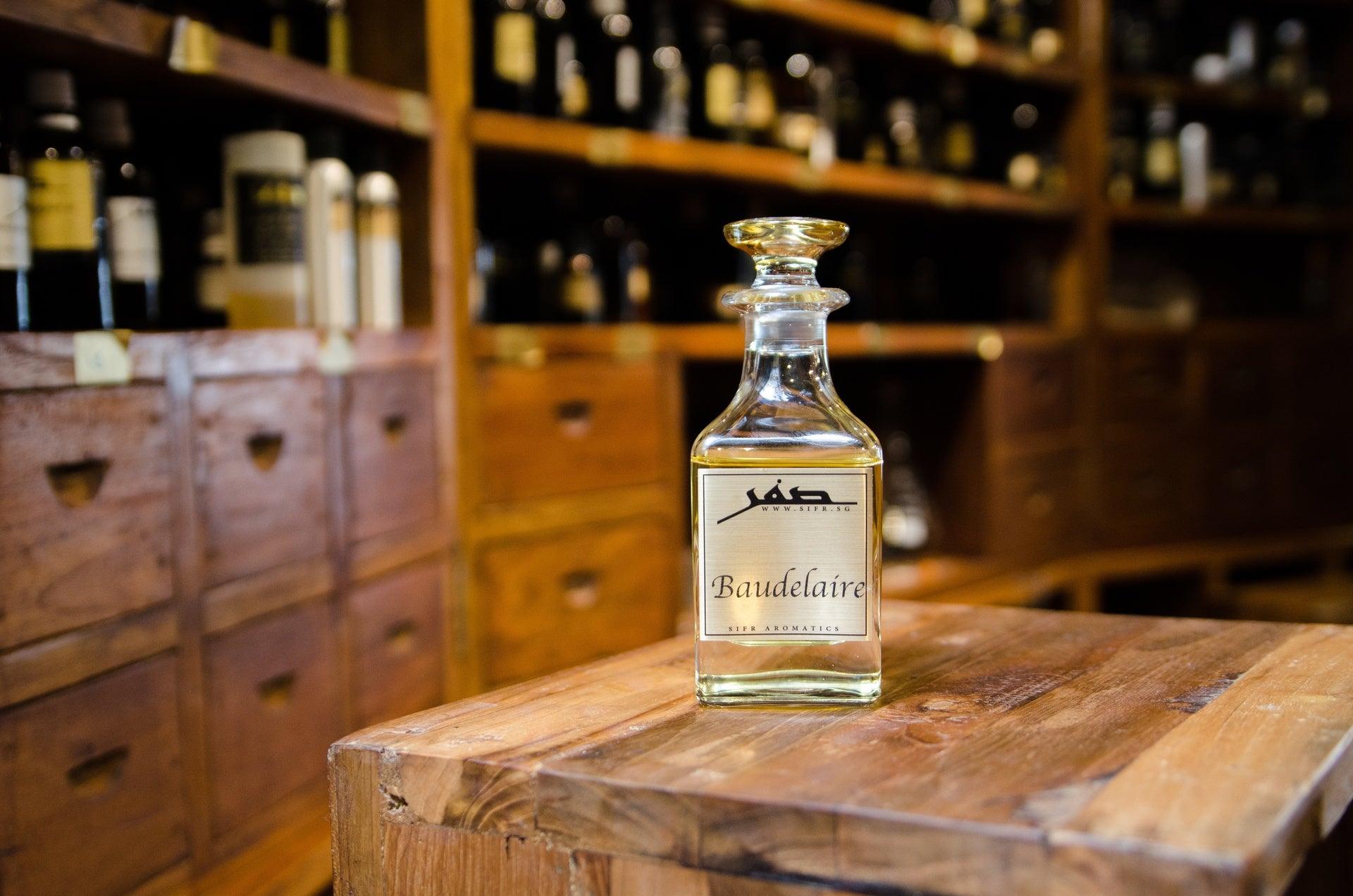 Ornate glass bottle for perfume oil with a gold label displaying the Sifr Aromatics logo and the name 'Baudelaire.' The background features an out-of-focus wooden perfume organ with various ingredients, creating an aesthetically pleasing scene