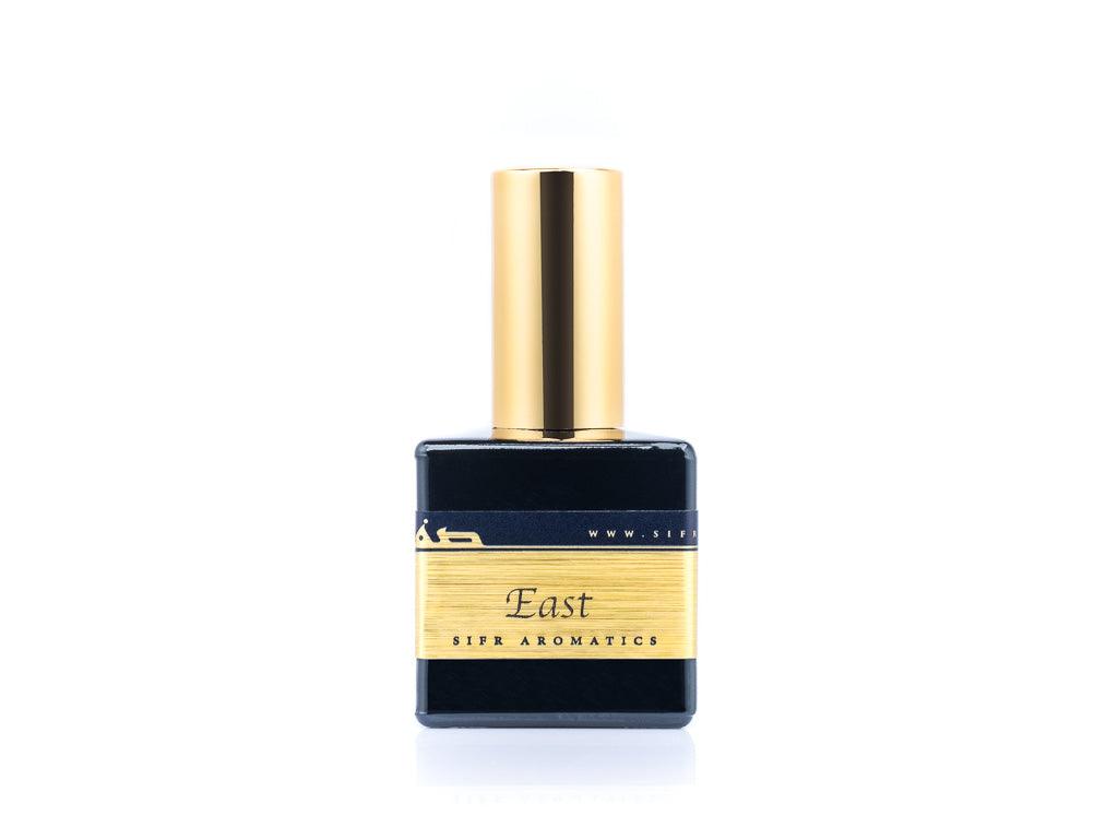 50ml spray bottle of Eau De Parfum placed elegantly on a white background. The bottle is sleek and black, with a polished gold cap. The label on the bottle is designed in gold and black, featuring Sifr Aromatics' logo and the perfume name 'East' The overall aesthetic is clean and sophisticated, highlighting the high-quality craftsmanship of the product